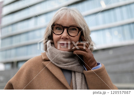 An elderly woman in a brown coat and scarf talks on her smartphone outside a modern building during An elderly woman in a brown coat and scarf talks on her smartphone outside a modern building during 120557087