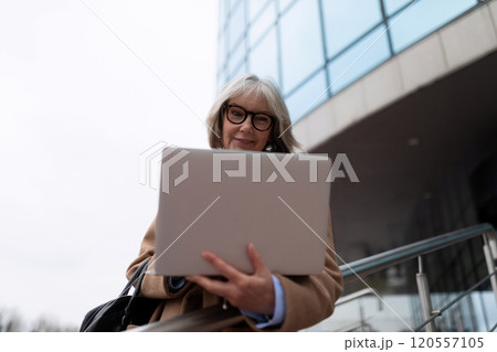 Senior woman using a laptop outdoors in front of a modern building during the day, showcasing work 120557105