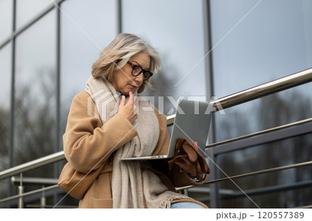 Older woman working on a laptop outdoors while seated on a railing in an urban environment during a Older woman working on a laptop outdoors while seated on a railing in an urban environment during a 120557389