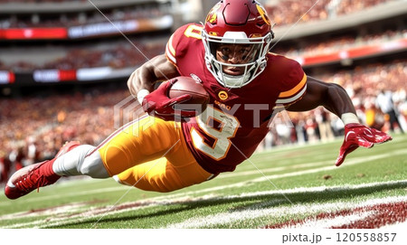 A football player is diving for the end zone, holding the ball in hand, wearing a red and gold uniform with a helmet, with a stadium background 120558857