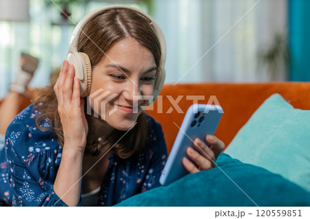 Young woman in wireless headphones relaxing lying on couch at home using smartphone listening music 120559851