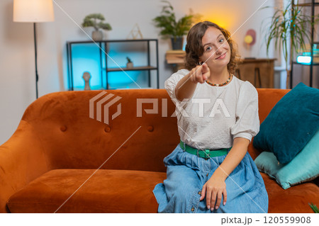 Young woman smiling and pointing to camera, choosing inviting, select sitting on sofa couch at home 120559908
