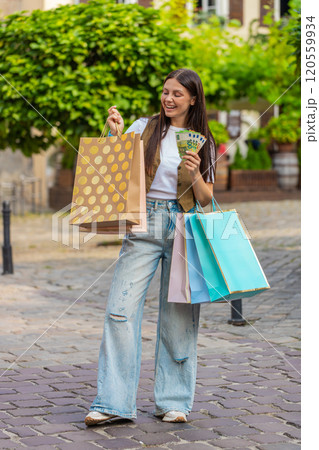Happy young shopaholic Caucasian woman holding euro cash while carrying shopping bags on city street 120559934