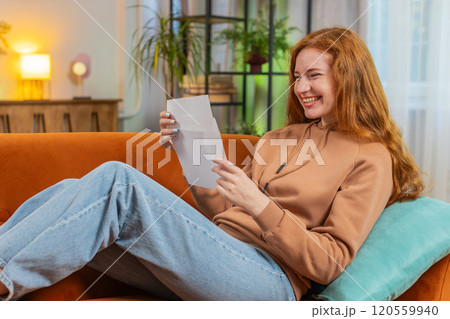 Excited young woman opening envelope letter and reading good news sitting on sofa at home apartment 120559940
