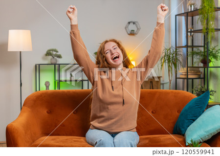 Happy young woman watching TV celebrating success, winning, lottery luck, victory sitting on sofa 120559941