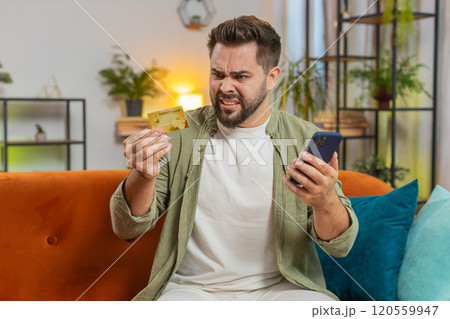 Angry stressed man trying to pay online shopping with smartphone blocked credit card on sofa at home Angry stressed man trying to pay online shopping with smartphone blocked credit card on sofa at home 120559947