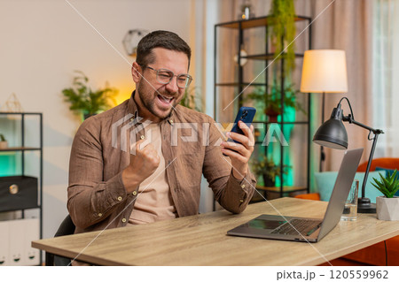 Happy young man freelancer using mobile phone celebrating success victory winning lottery at table 120559962