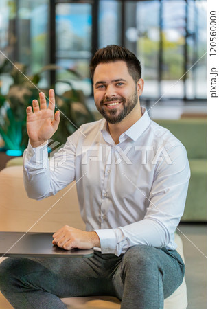 Portrait of smiling young businessman with laptop wave hand at camera sitting in hotel lobby 120560000