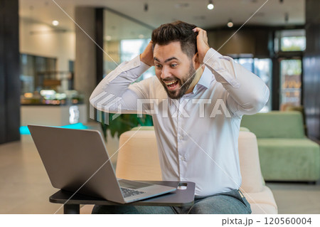 Caucasian bearded businessman working on laptop shocked by sudden victory in modern hotel lobby 120560004