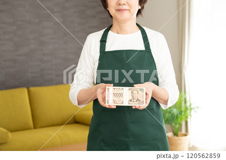100万円の札束を持って微笑む緑のエプロンをしたミドル〜シニア女性の主婦 120562859