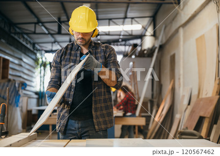 Construction worker in a yellow hardhat and safety glasses inspecting a board of wood. Ideal for carpentry, woodworking, and quality control, highlighting craftsmanship in the workshop. Labor day 120567462