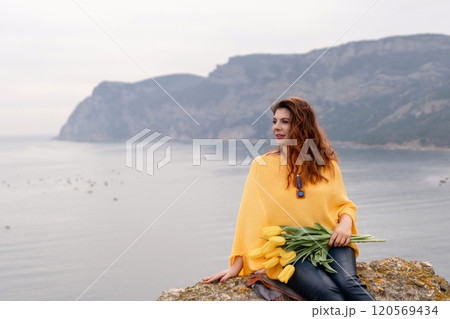 Portrait happy woman woman with long hair against a background of mountains and sea. Holding a bouquet of yellow tulips in her hands, wearing a yellow sweater Portrait happy woman woman with long hair against a background of mountains and sea. Holding a bouquet of yellow tulips in her hands, wearing a yellow sweater 120569434