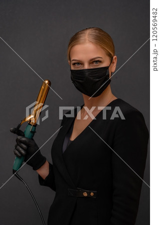 A woman in a black dress holding a hair curler. She is wearing a mask. Concept of caution and responsibility in the face of a pandemic. A woman in a black dress holding a hair curler. She is wearing a mask. Concept of caution and responsibility in the face of a pandemic. 120569482