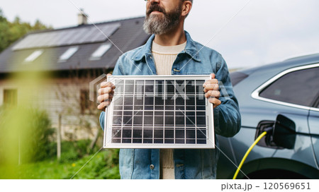 Man holding model of solar panel in hands. Charging electric car and house with solar panel system on roof behind him. 120569651