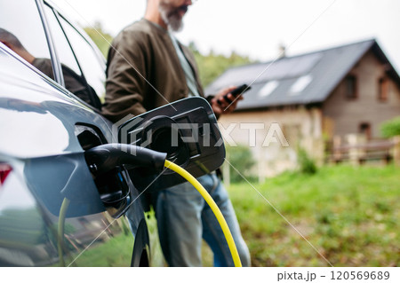 Close up of charging electric car in front of house, charger in charging port. House with solar panel system on roof behind. 120569689