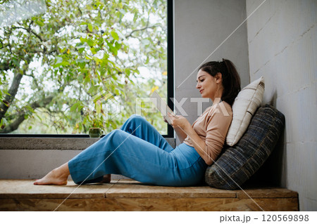Hygge moment for beautiful woman sitting by a large window with a book. Woman is enjoying time for herself, reading. 120569898