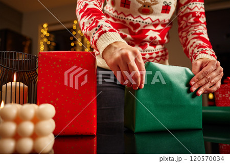 Female hands wrapping Christmas gifts in red and green paper. Woman in festive sweater preparing presents for winter holidays. Christmas and Happy New Year family traditions 120570034
