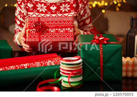 Woman wearing Christmas sweater holding wrapped Christmas gift with wrapping paper and ribbon table. Preparing presents for winter holidays 120570055