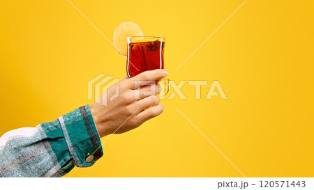 Woman's hand holds glass of hot mulled wine, garnished with apple slice and cinnamon stick, against warm yellow studio background. Woman's hand holds glass of hot mulled wine, garnished with apple slice and cinnamon stick, against warm yellow studio background. 120571443