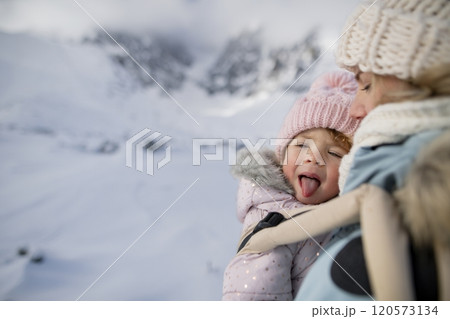 Mother standing in the middle of snowy landscape with daughter in baby carrier. Mom and girl enjoying winter holiday in the mountains. 120573134