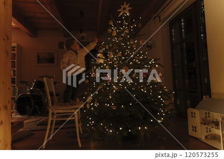 Portrait of cute boy standing on chair and helping with decorating the Christmas tree with ornaments. Portrait of cute boy standing on chair and helping with decorating the Christmas tree with ornaments. 120573255