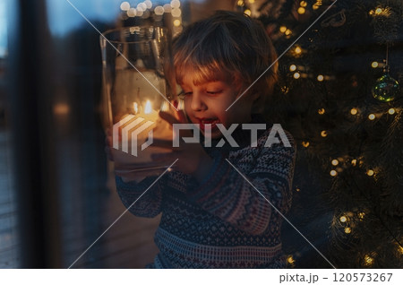 Cute little boy holding Christmas candle in his hands, Christmas tree behind him. Festive atmosphere at home. 120573267