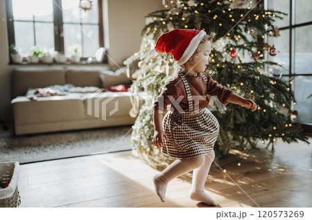 Portrait of cute girl in Santa's hat helping with decorating the Christmas tree and dancing around the tree. 120573269