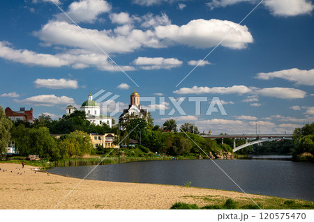 A quiet riverside location with a sandy beach, lush greenery and historic architecture with churches and cathedrals. The bridge spans the river under a bright blue sky with fluffy white clouds. 120575470