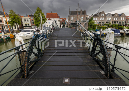 A dark, rainy bridge leads towards a charming marina with boats and historic buildings under a dramatic, stormy sky. Wet wooden planks and intricate ironwork add to the atmosphere. A dark, rainy bridge leads towards a charming marina with boats and historic buildings under a dramatic, stormy sky. Wet wooden planks and intricate ironwork add to the atmosphere. 120575472