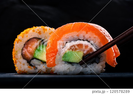 Two sushi rolls with salmon and avocado, on a black background with chopsticks 120578289