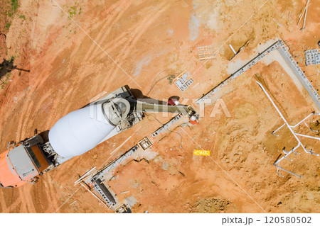 Worker pouring concrete along foundations in spacious area with reddish soil using concrete mixer truck. 120580502