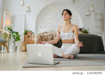 Yoga mindfulness meditation online. Woman practicing yoga with online lessons in laptop at home. Woman sitting in lotus pose meditating relaxing indoor. Girl doing breathing practice. Yoga at home 120586895