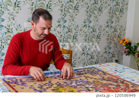 Bearded hispanic man in his 40s wearing a red sweater enjoying his free time while having fun doing a puzzle in the dining room of his house. Bearded hispanic man in his 40s wearing a red sweater enjoying his free time while having fun doing a puzzle in the dining room of his house. 120586949