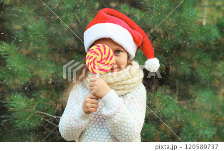 Christmas portrait of happy smiling little girl child in santa red hat with sweet lollipop outdoors Christmas portrait of happy smiling little girl child in santa red hat with sweet lollipop outdoors 120589737