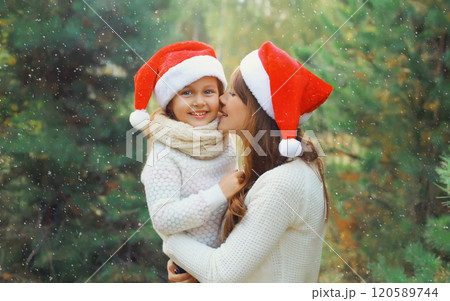 Family, holiday. Happy mother kissing her little girl child in santa hat near Christmas tree outdoor 120589744