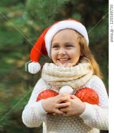 Christmas portrait of happy smiling little girl child in santa red hat with ball decoration outdoors 120589750