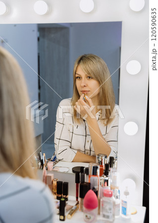 Reflection of a woman applying lipstick in a makeup mirror with beauty products on the table 120590635