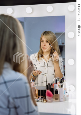 Woman applying makeup in front of a vanity mirror with bright lights for a flawless look 120590644