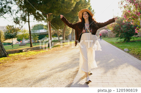 A Joyful Woman is Walking in Nature, Dressed in a Flowing Outfit and Wearing a Sun Hat 120590819