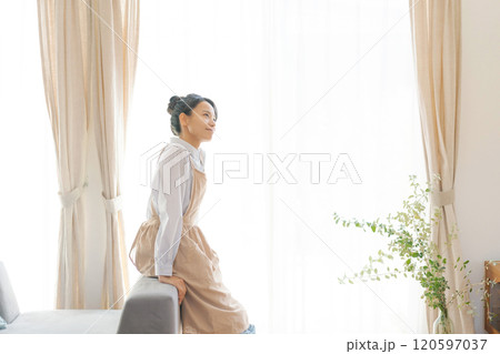 リビングの窓際に立つエプロン姿の女性 120597037