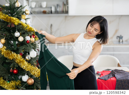 Sad young Asian girl trying to choose a dress standing next to Xmas tree Sad young Asian girl trying to choose a dress standing next to Xmas tree 120598342