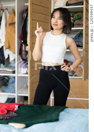 Happy young Asian girl trying to choose a dress standing by wardrobe 120598437