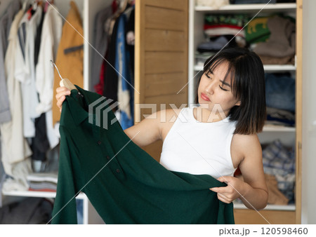 Sad young Asian girl trying to choose a dress standing by wardrobe 120598460