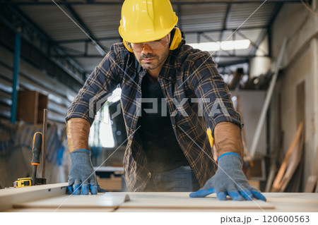 Young carpenter in a yellow helmet, blue gloves, and plaid shirt using a circular saw to cut wood in a furniture factory. Ideal for showcasing woodworking skills and construction techniques. Labor day Young carpenter in a yellow helmet, blue gloves, and plaid shirt using a circular saw to cut wood in a furniture factory. Ideal for showcasing woodworking skills and construction techniques. Labor day 120600563