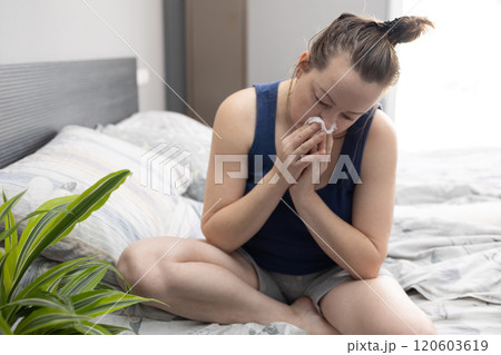 A woman is sitting comfortably on a bed while blowing her nose into a napkin 120603619