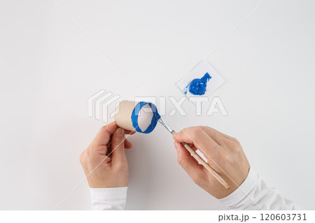 a person painting the edge of a cardboard tube with blue paint using a paintbrush. step in a process of crafting or DIY activity, creative use of everyday materials like a cardboard tube and paint 120603731