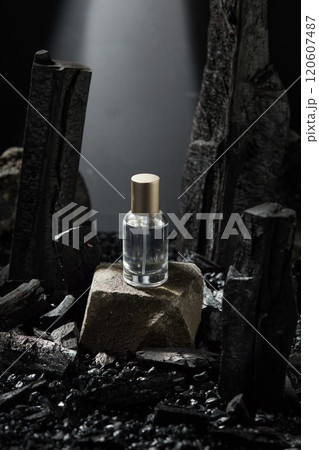 An advertisement photo template in the dark background, high angle, shows a glass bottle containing a transparent liquid placed on a stone block. Charcoal ingredient and gravels surrounded a surface. 120607487