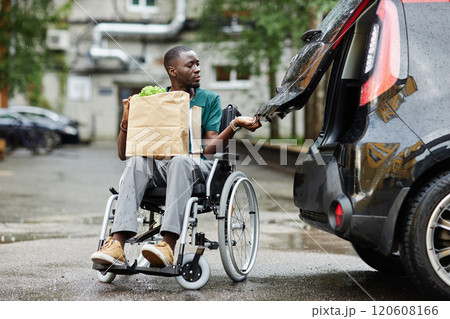 Full length scene with of young African American man using wheelchair opening car trunk in parking lot and holding groceries while doing shopping in city 120608166