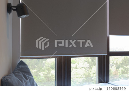 Roller blinds motorized. Automatic roller shades beige color on large windows. Remote control curtains are near the sofa with the pillow. Sofa covered with pastel velours. Summer. Green trees outside. 120608569