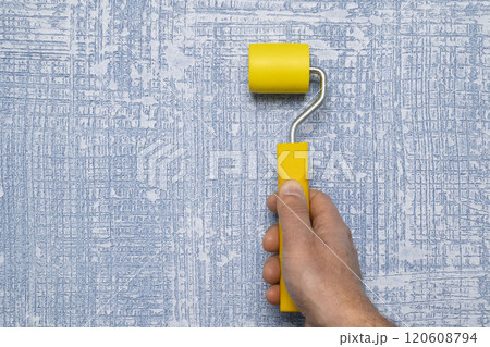 A persons hand is skillfully holding a bright yellow paint roller against a textured blue wall 120608794